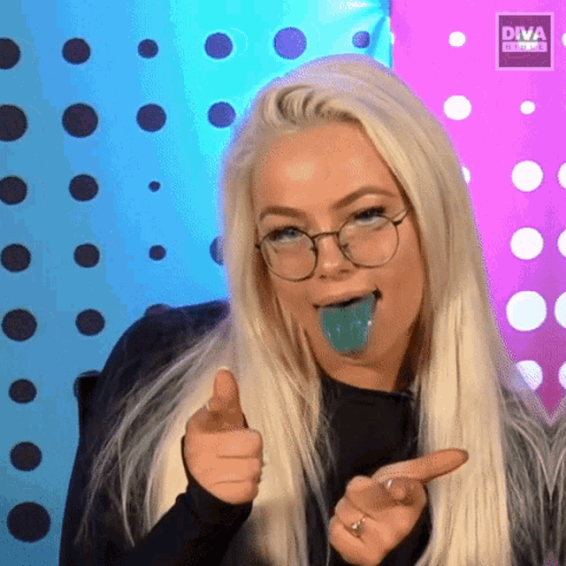 a blonde woman with glasses and a blue lollipop in her mouth is pointing at the camera and giving a thumbs up .