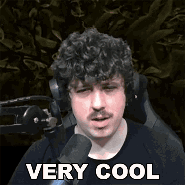 a man with a mustache is sitting in front of a microphone with the words very cool above him