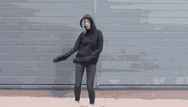 a woman in a black hoodie and jeans is dancing in front of a gray wall .
