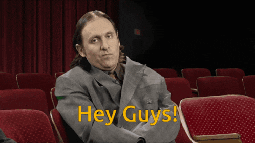 a man in a suit sits in a theater with the words " hey guys " written in yellow