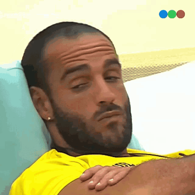 a man with a beard wearing a yellow shirt is laying on a bed with his eyes closed