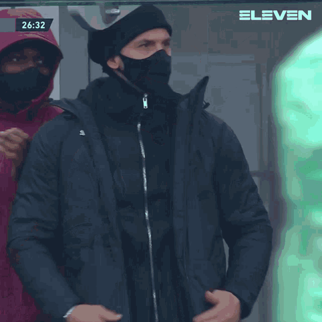 a man wearing a mask and a beanie stands in front of a screen that says 6:32