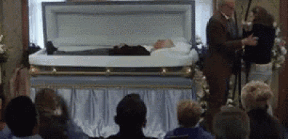 a group of people are sitting in front of a coffin with a man standing behind it .