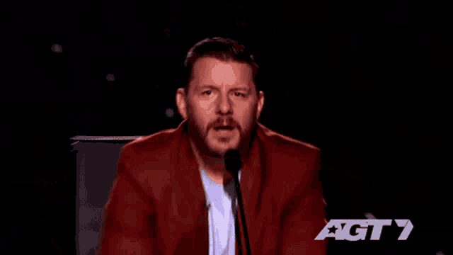 a man in a red suit is speaking into a microphone with the letters agt 7 behind him