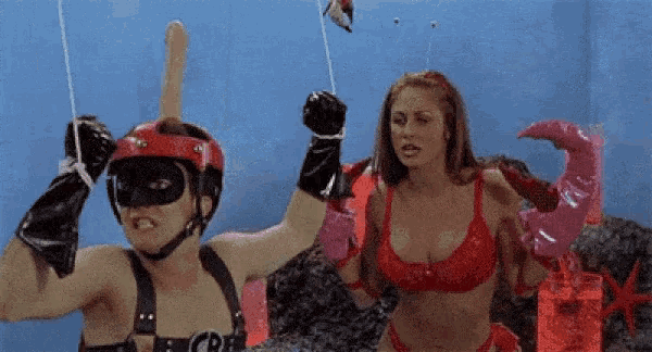a man in a mask and gloves is holding a dildo while a woman in a red bra stands behind him