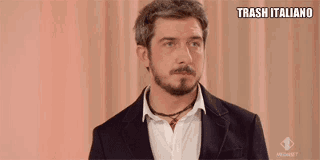 a man with a beard is wearing a black jacket and a white shirt and says trash italiano on the bottom right