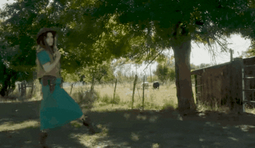 a woman in a blue dress and cowboy hat is standing under a tree .