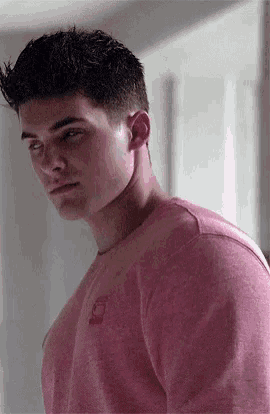 a young man wearing a pink shirt is standing in front of a door and looking at the camera .
