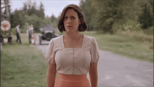 a woman in a white top and pink skirt stands on a dirt road