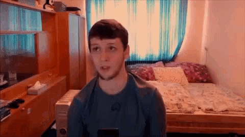 a young man is sitting in front of a bed in a bedroom looking at the camera .