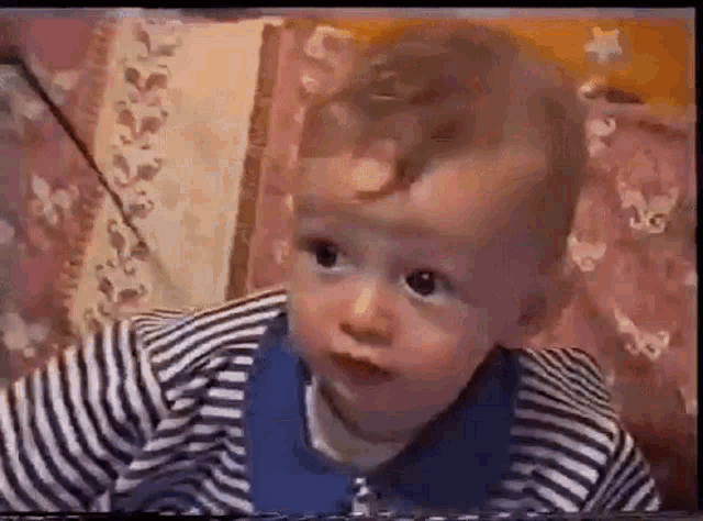 a baby in a striped shirt is sitting on a bed looking at the camera .
