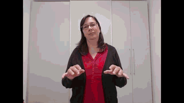 a woman wearing glasses and a red shirt is standing in front of a white wall and making a gesture with her hands .