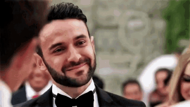 a man with a beard is wearing a tuxedo and bow tie and smiling at another man .