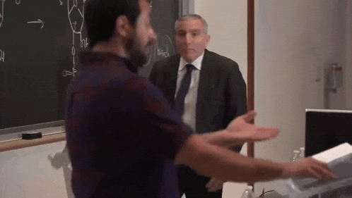 a man in a suit and tie is standing next to another man in a classroom .