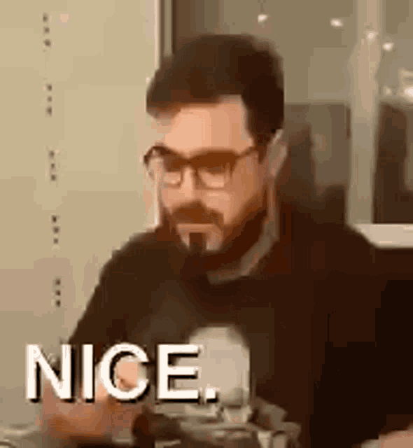 a man with glasses and a beard is sitting at a table with the words `` nice '' written on the screen .