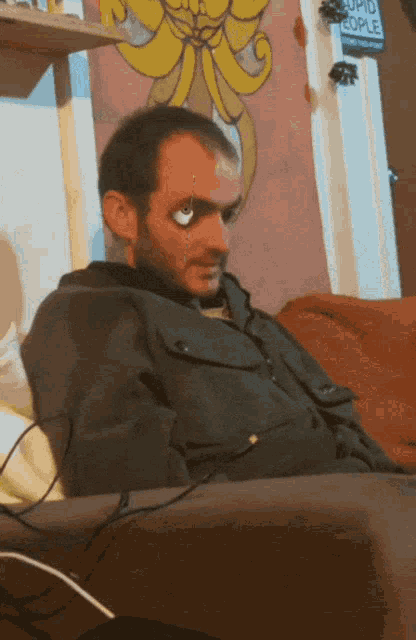 a man is sitting on a couch in front of a wall with a sticker that says stupid people
