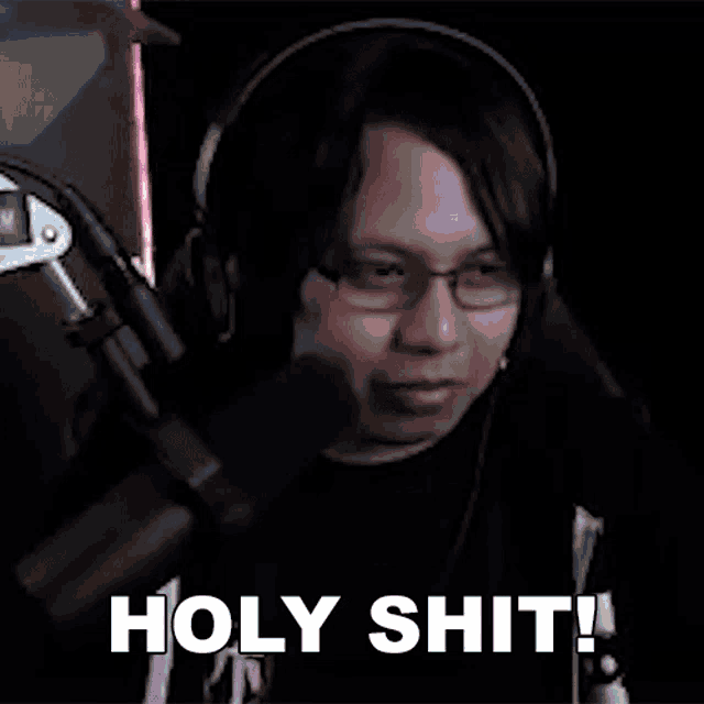 a man wearing headphones and glasses is sitting in front of a microphone and saying holy shit !