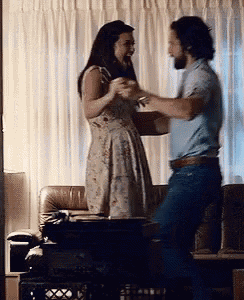 a man and a woman are dancing in a living room . the woman is wearing a dress and the man is wearing jeans .