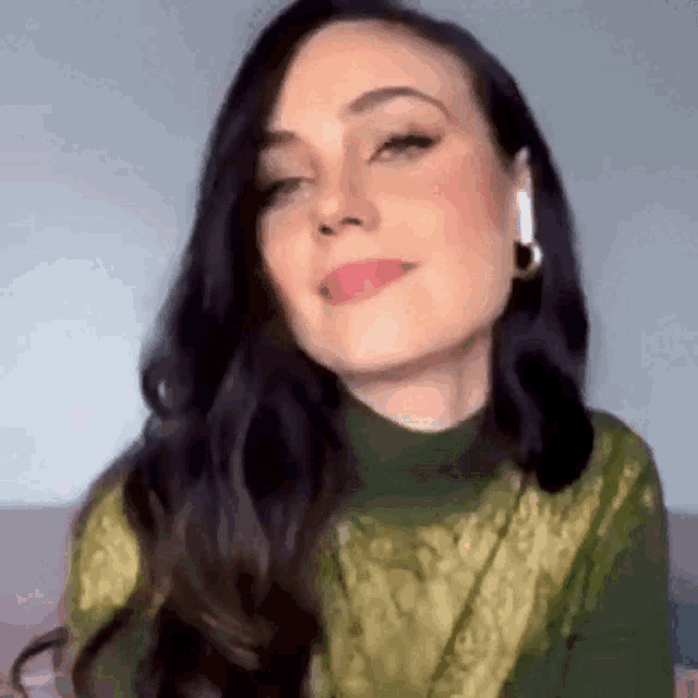 a woman with long dark hair is wearing a green sweater and earrings and looking at the camera .