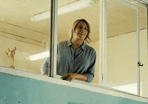 a woman in a blue shirt is smiling while looking out of a window .