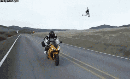 a man is riding a motorcycle down a road while a person on a hoverboard is flying overhead .