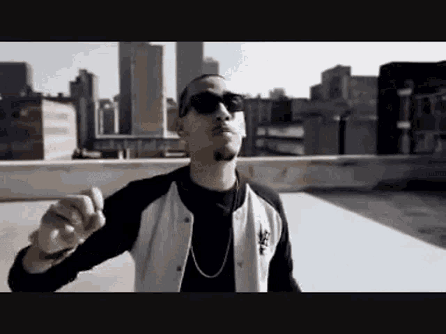 a man wearing sunglasses and a white jacket is standing on a rooftop in front of a city .