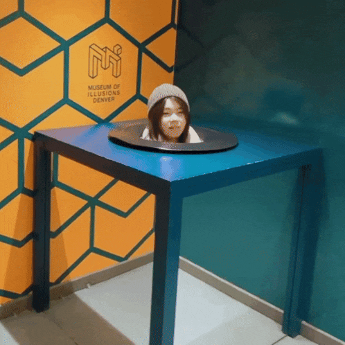 a woman 's head is sticking out of a blue table at the museum of illusions in denver