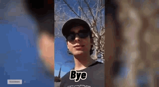 a young man wearing sunglasses and a baseball cap is standing in front of a tree .