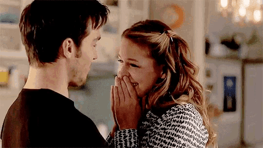 a man and a woman are laughing together in a kitchen . the woman is covering her mouth with her hand .