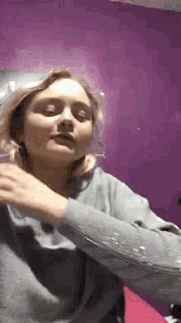 a woman in a gray sweater is standing in front of a purple wall and making a funny face .