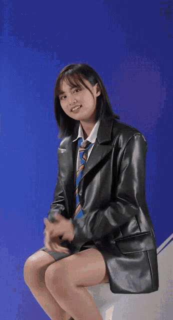 a woman wearing a leather jacket and tie is sitting on a stool