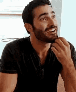 a man with a beard is sitting at a table with his hand on his chin and smiling .