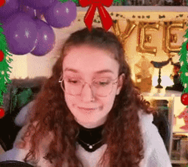 a woman wearing glasses and a bow on her head is sitting in front of balloons and a christmas tree .