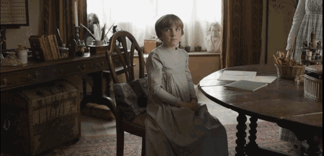 a little girl in a white dress sits at a table with papers on it