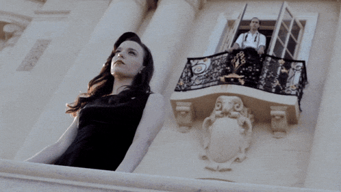 a woman in a black dress stands on a balcony looking at a man