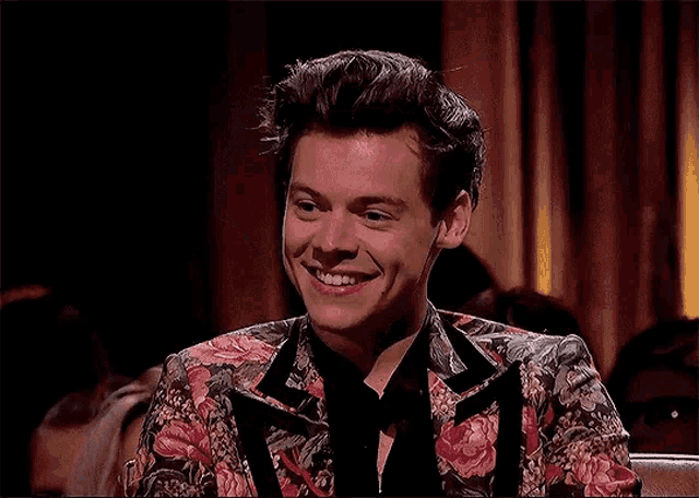 harry styles is wearing a floral suit and sitting at a table with his hand up .