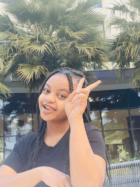 a girl with braids giving the peace sign in front of a palm tree
