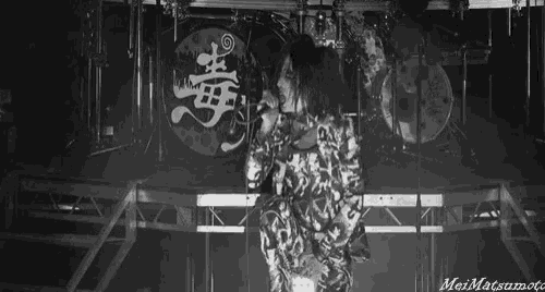 a black and white photo of a person singing in front of a drum set and a sign that says meimatsumoto