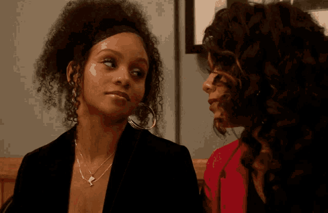 two women looking at each other with one wearing a necklace with a letter k on it