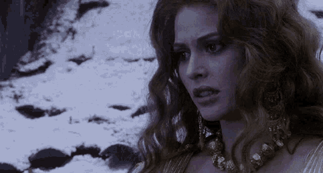 a woman with long hair and a gold necklace is standing in the snow looking at the camera .