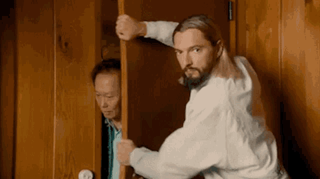 a man with long hair and a beard is holding a door open while another man looks out .