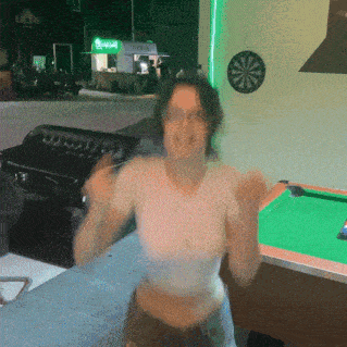 a woman stands in front of a pool table with a dart board on the wall above her