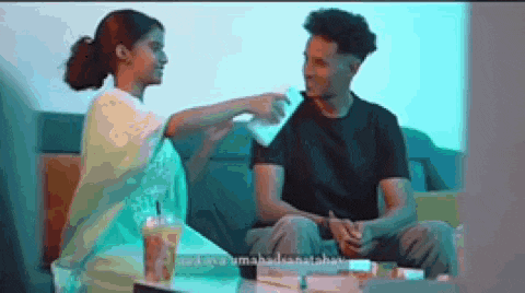 a man and a woman are sitting on a couch and the woman is feeding the man a drink .