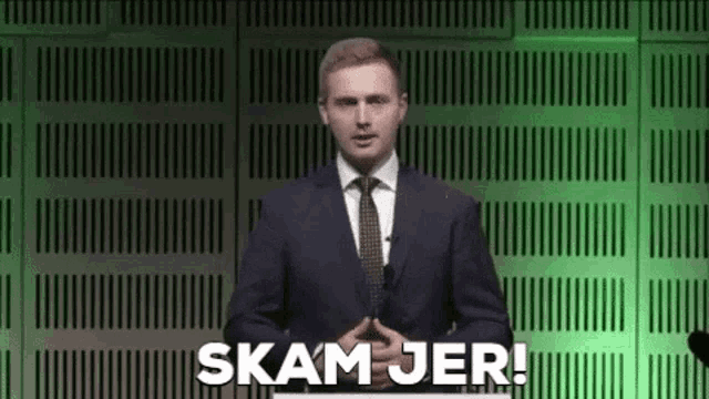 a man in a suit and tie is standing in front of a green wall and giving a presentation .