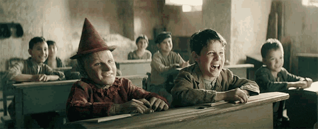 a group of children are sitting at desks in a classroom and one of them is wearing a witch hat