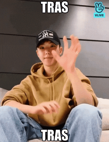 a young man wearing a ny hat is sitting on a couch and making a hand gesture with the word tras below him
