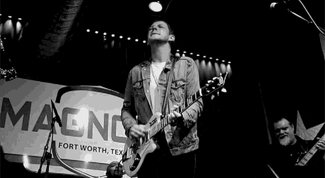 a black and white photo of a man playing a guitar in front of a sign that says magnolia fort worth texas