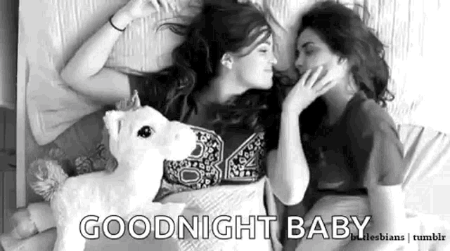 two women are laying on a bed with a stuffed animal unicorn .