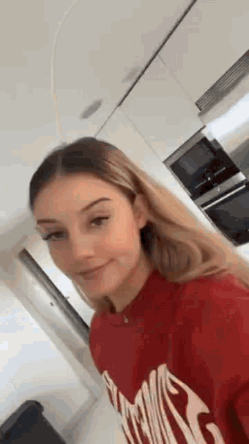 a woman is taking a selfie in a kitchen wearing a red sweatshirt .