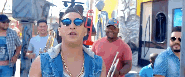 a man wearing sunglasses and a denim vest is standing in front of a group of men playing instruments .